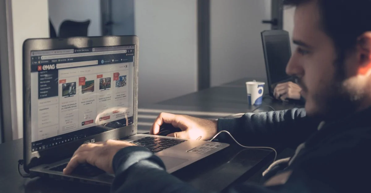 Homme adulte utilisant un ordinateur portable pour naviguer sur un marketplace en ligne dans un bureau moderne.