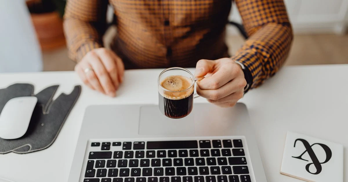 Une personne tenant un espresso près d'un ordinateur portable et d'une souris, idéal pour des thèmes de travail à domicile.