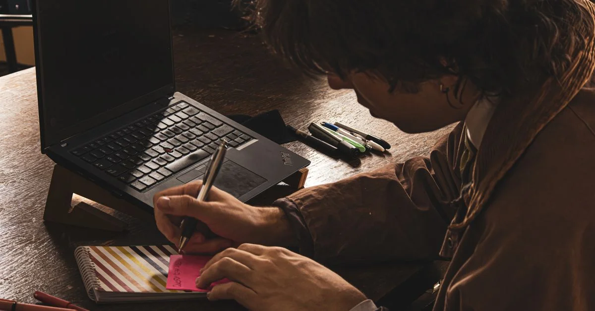 Un jeune homme prend des notes au stylo à côté d'un ordinateur portable dans un bureau faiblement éclairé.
