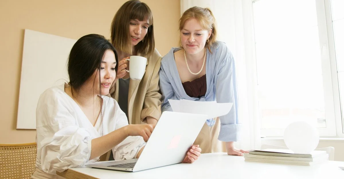 Trois femmes dans un bureau moderne collaborant autour d'un ordinateur portable et discutant de documents.