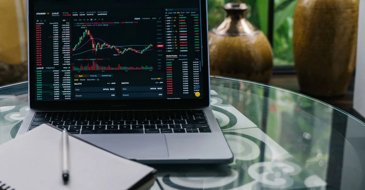 Ordinateur portable affichant des actions et des graphiques de cryptomonnaies sur une table en verre avec un carnet.