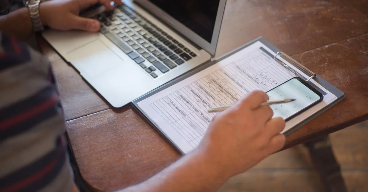 Gros plan sur une personne utilisant un ordinateur portable et consultant des documents avec un smartphone sur une table en bois.