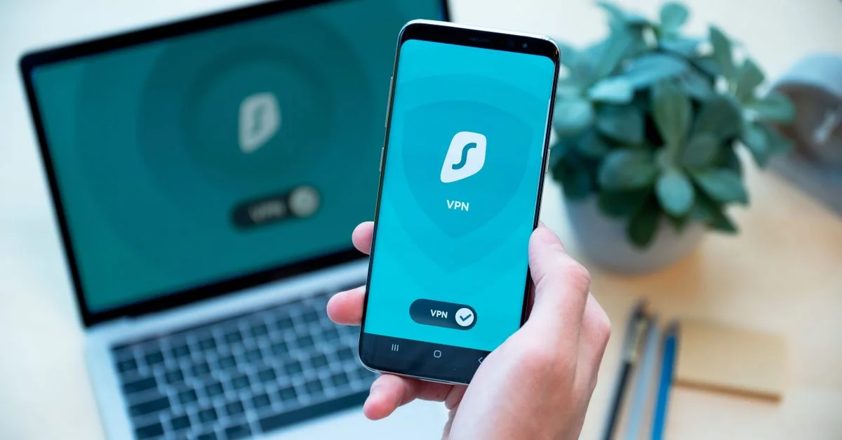 Gros plan sur une main tenant un smartphone avec une application VPN, ordinateur portable en arrière-plan, mettant en avant la sécurité numérique.